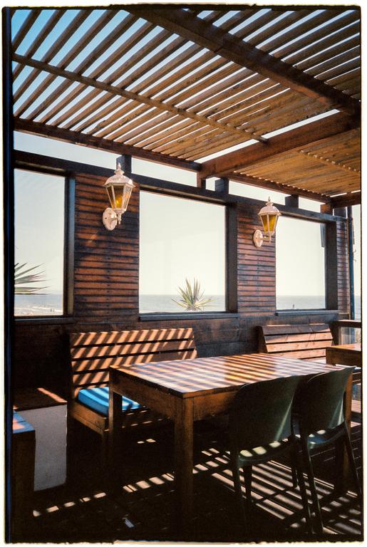 A wooden patio with a slatted roof and furniture is illuminated by strong shadows, with the ocean visible through windows in the background.