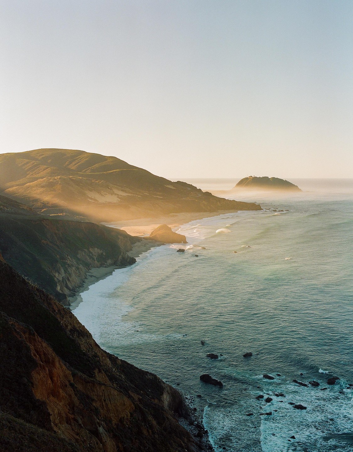 Sunlight illuminates a mountainous coastline with waves crashing on the shore.