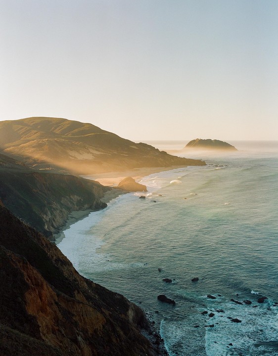 Sunlight illuminates a mountainous coastline with waves crashing on the shore.