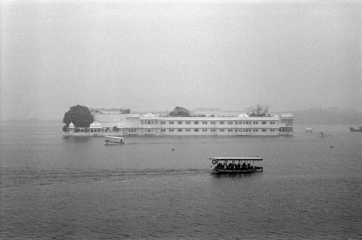 A large white building with many windows sits on an island in the middle of a body of water, with several boats moving across the water.