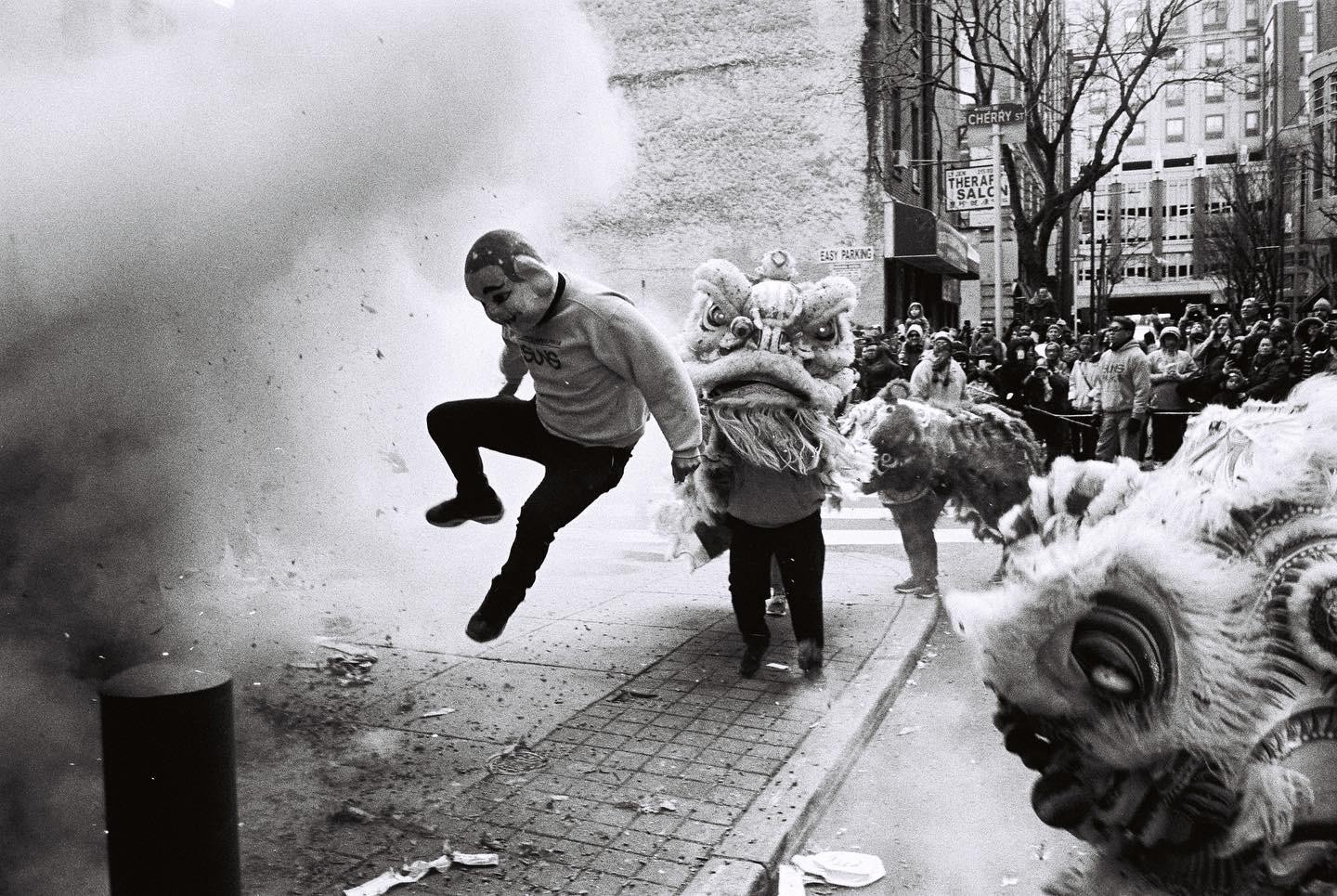 A man in a mask jumps away from an explosion of smoke and debris next to two lion dancers on a city street.