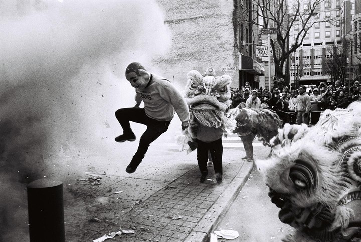 A man in a mask jumps away from an explosion of smoke and debris next to two lion dancers on a city street.