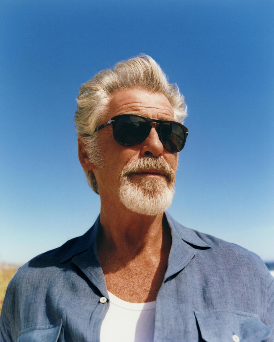 A man with gray hair and a beard wears sunglasses and a blue shirt against a clear blue sky.