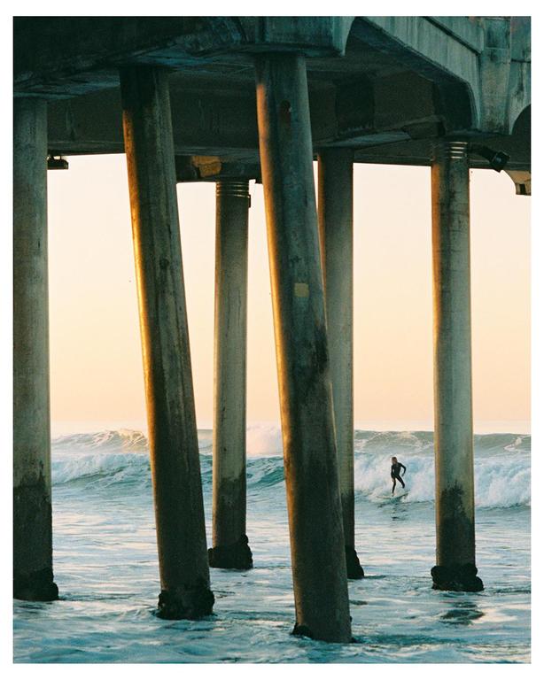 A surfer rides a wave between the pillars of a pier at golden hour.