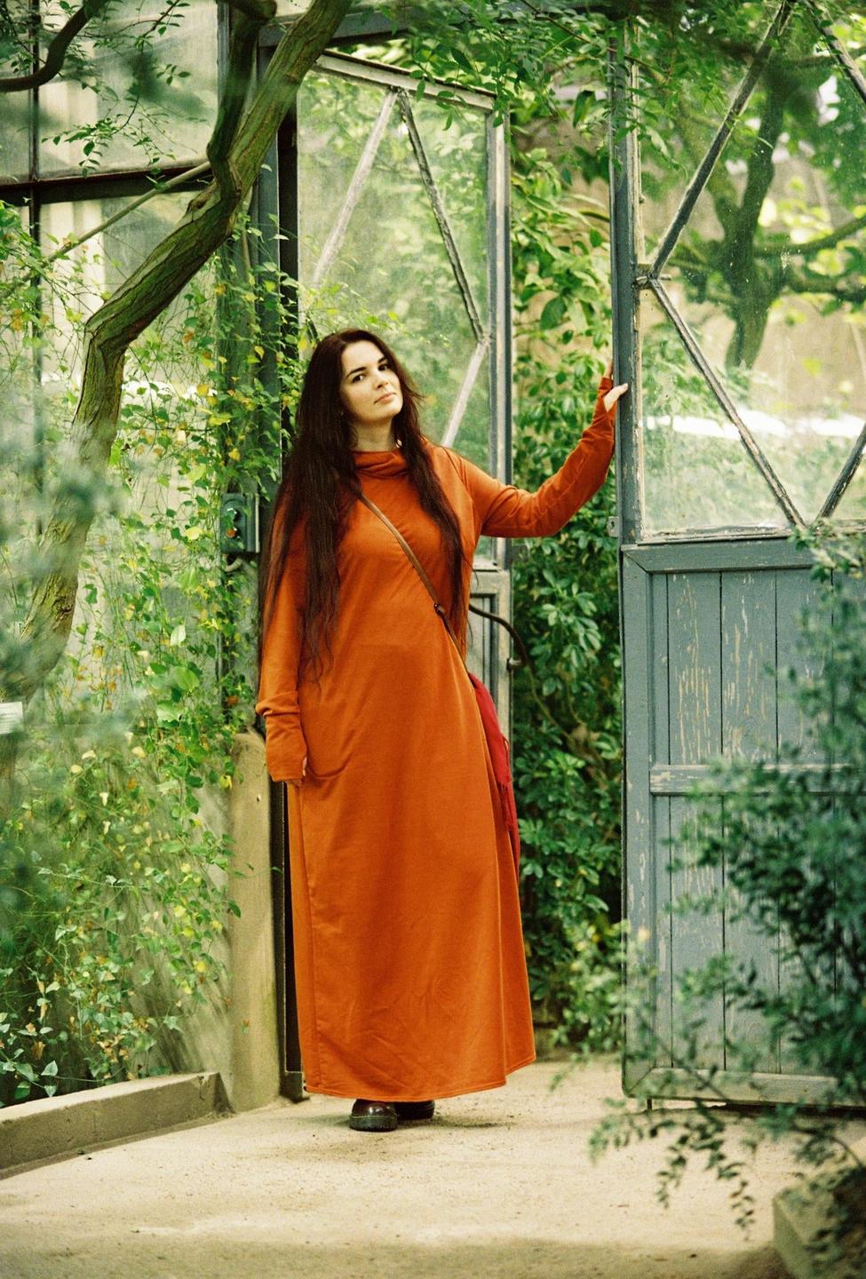 A woman in an orange dress stands in a greenhouse with lush green foliage.