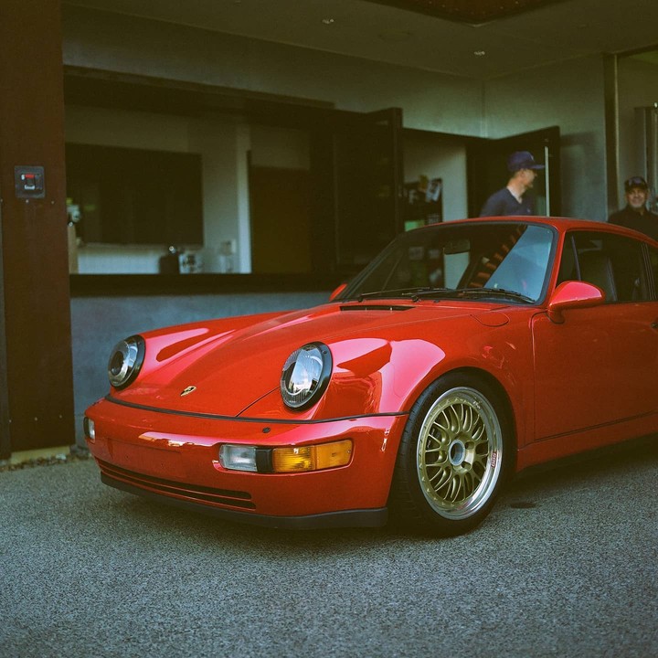 A red car with gold wheels is parked on asphalt with two men in the background.