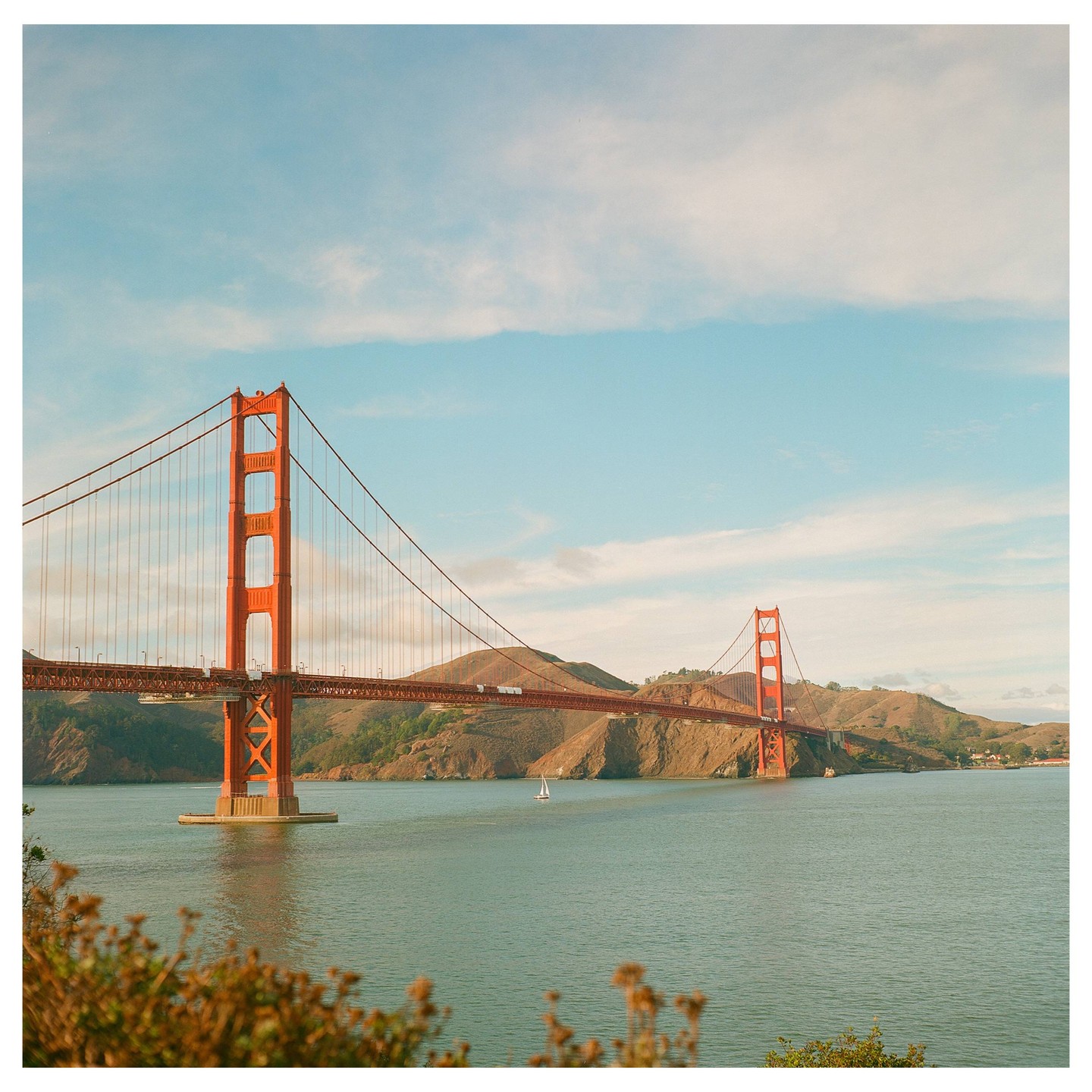 The Golden Gate Bridge spans a body of water with hills in the background under a partly cloudy sky.