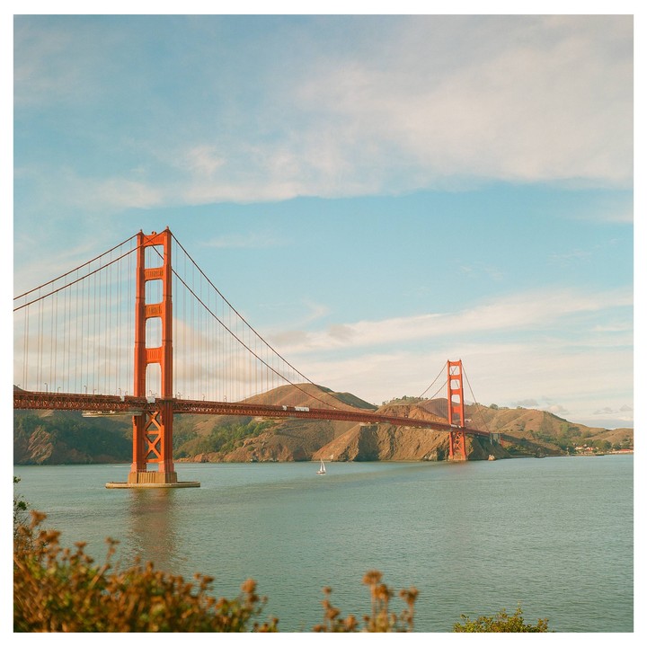 The Golden Gate Bridge spans a body of water with hills in the background under a partly cloudy sky.