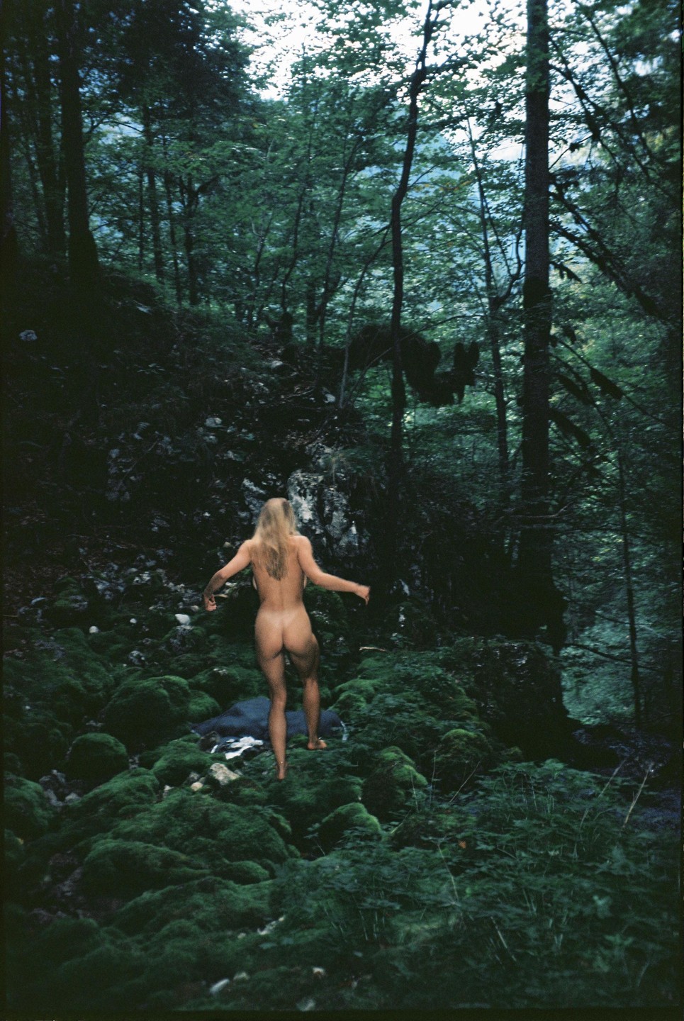 A nude woman with long blonde hair walks through a mossy forest.