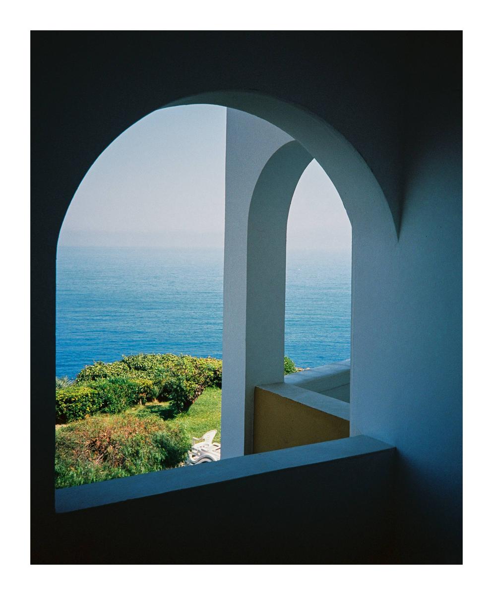A view of the sea and green foliage is framed by two white arches.