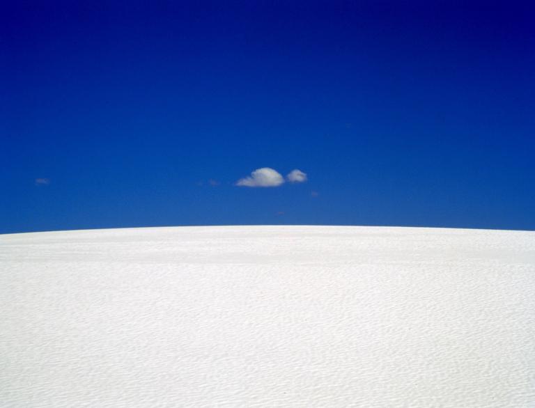 A small cloud floats in a deep blue sky above a white sand dune.