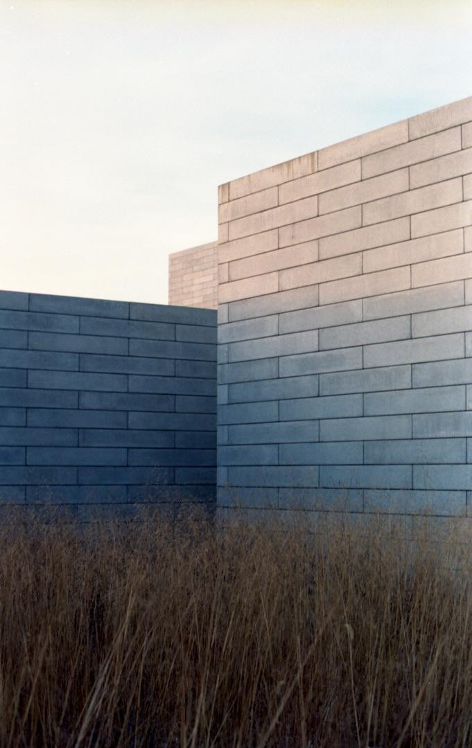 Three concrete walls rise from a field of dry grass under a pale sky.