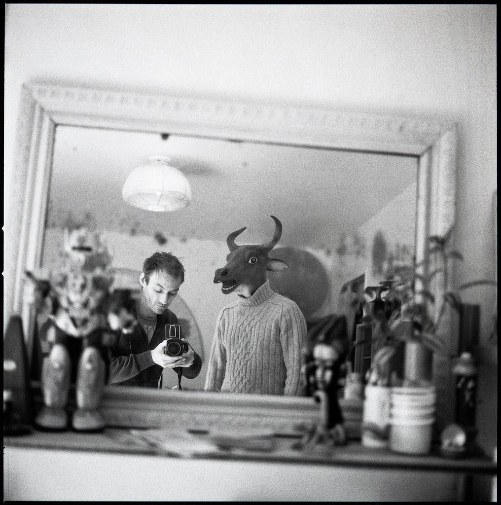 A man takes a mirror self-portrait next to a person wearing a cow mask and a sweater.