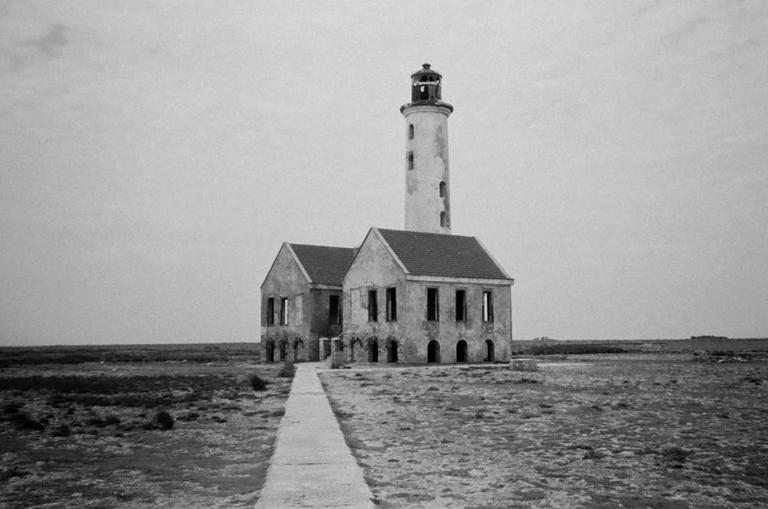 A tall lighthouse stands next to two abandoned buildings with a path leading towards them under a cloudy sky.