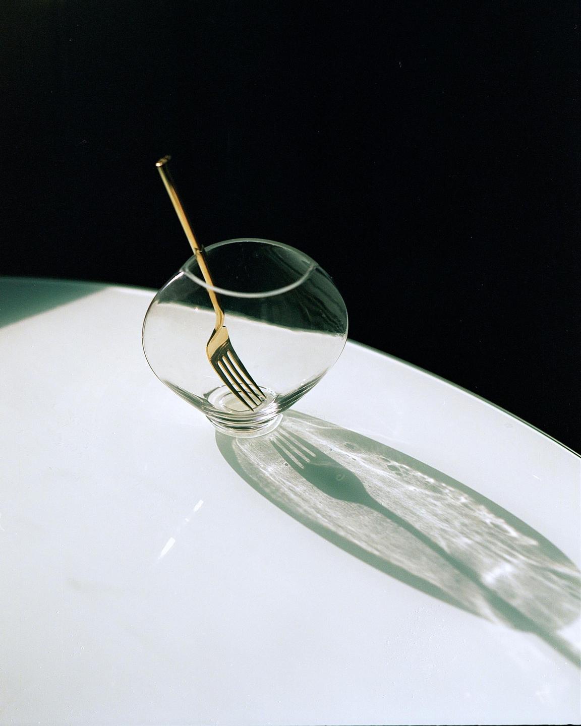 A golden fork is placed inside a clear glass bowl, casting a shadow on a white surface.