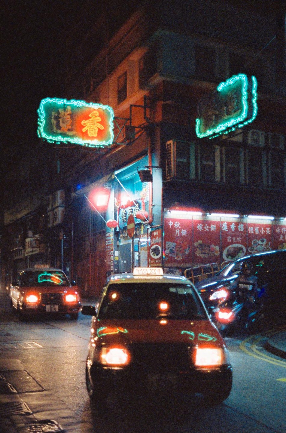 Two taxis drive down a street at night, illuminated by neon signs and storefront lights.