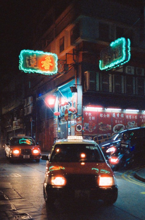 Two taxis drive down a street at night, illuminated by neon signs and storefront lights.