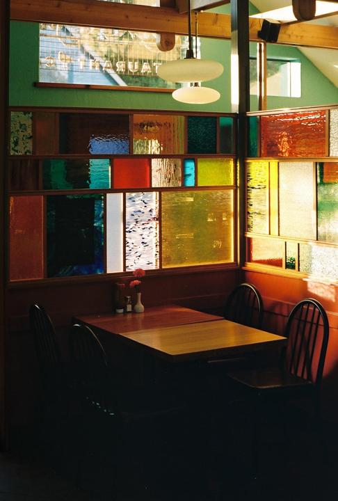 A restaurant booth with a wooden table and chairs is illuminated by light filtering through colorful stained glass panels.