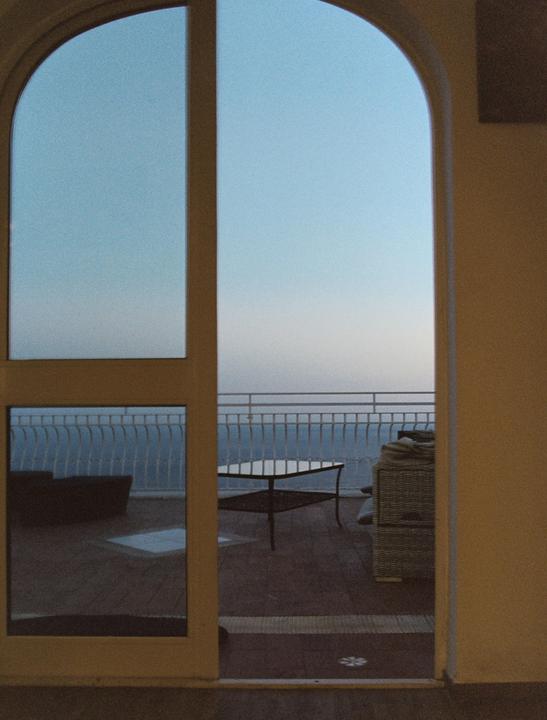 A view of a balcony with a table and chairs overlooking the ocean is seen through an arched doorway.