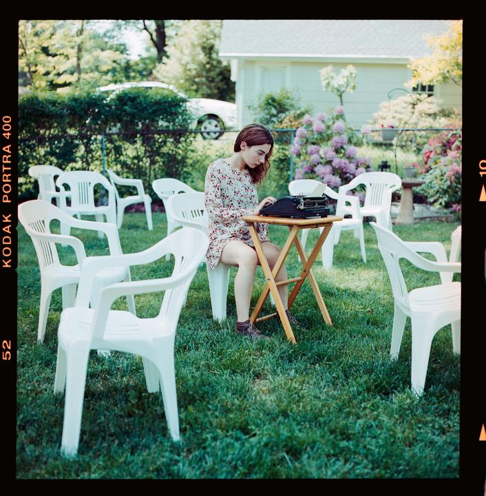 A woman sits at a small table with a typewriter in a grassy backyard surrounded by white plastic chairs.