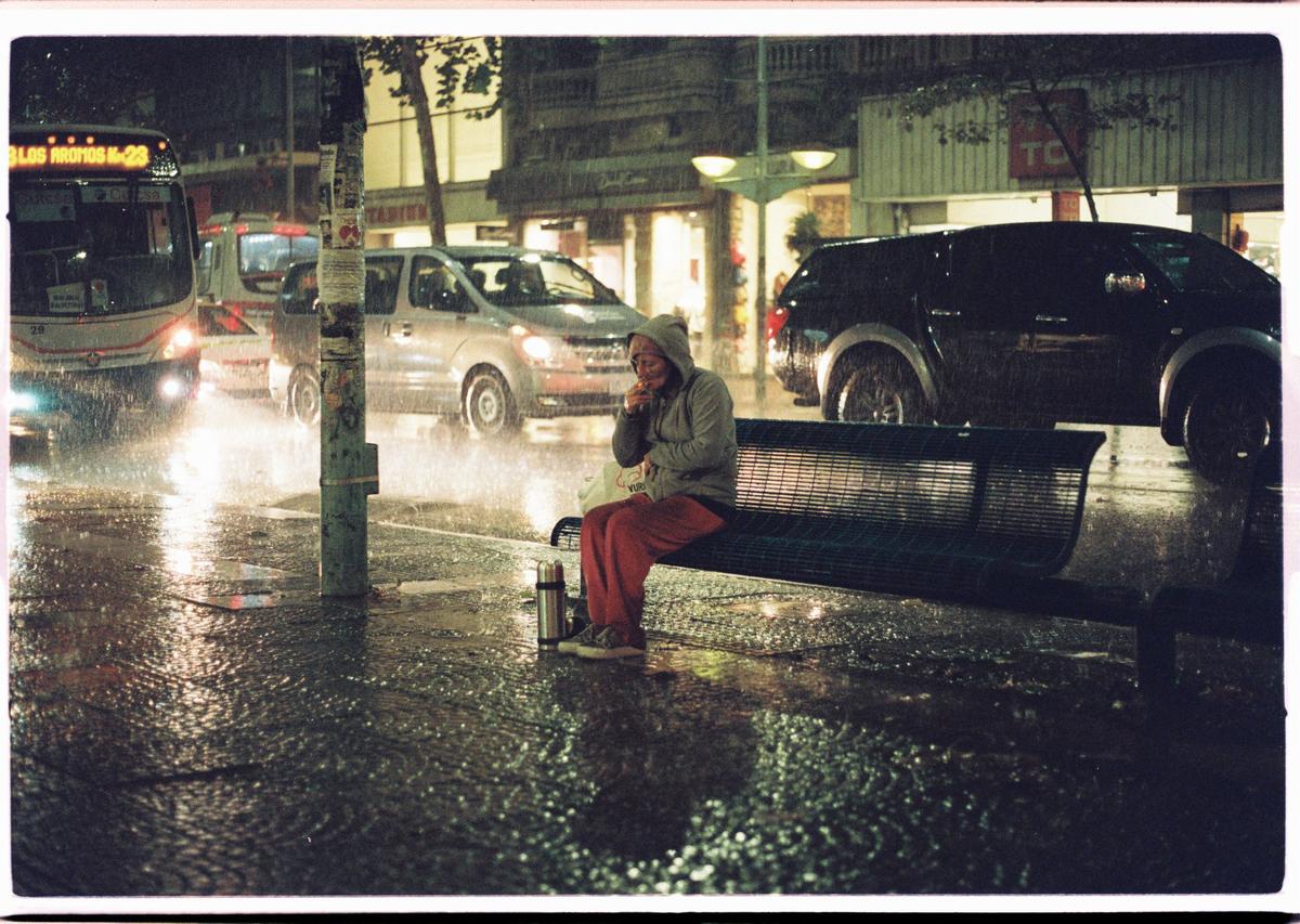 A person in a hooded sweatshirt sits on a bench in the rain at night, with cars and a bus passing by on a wet street.