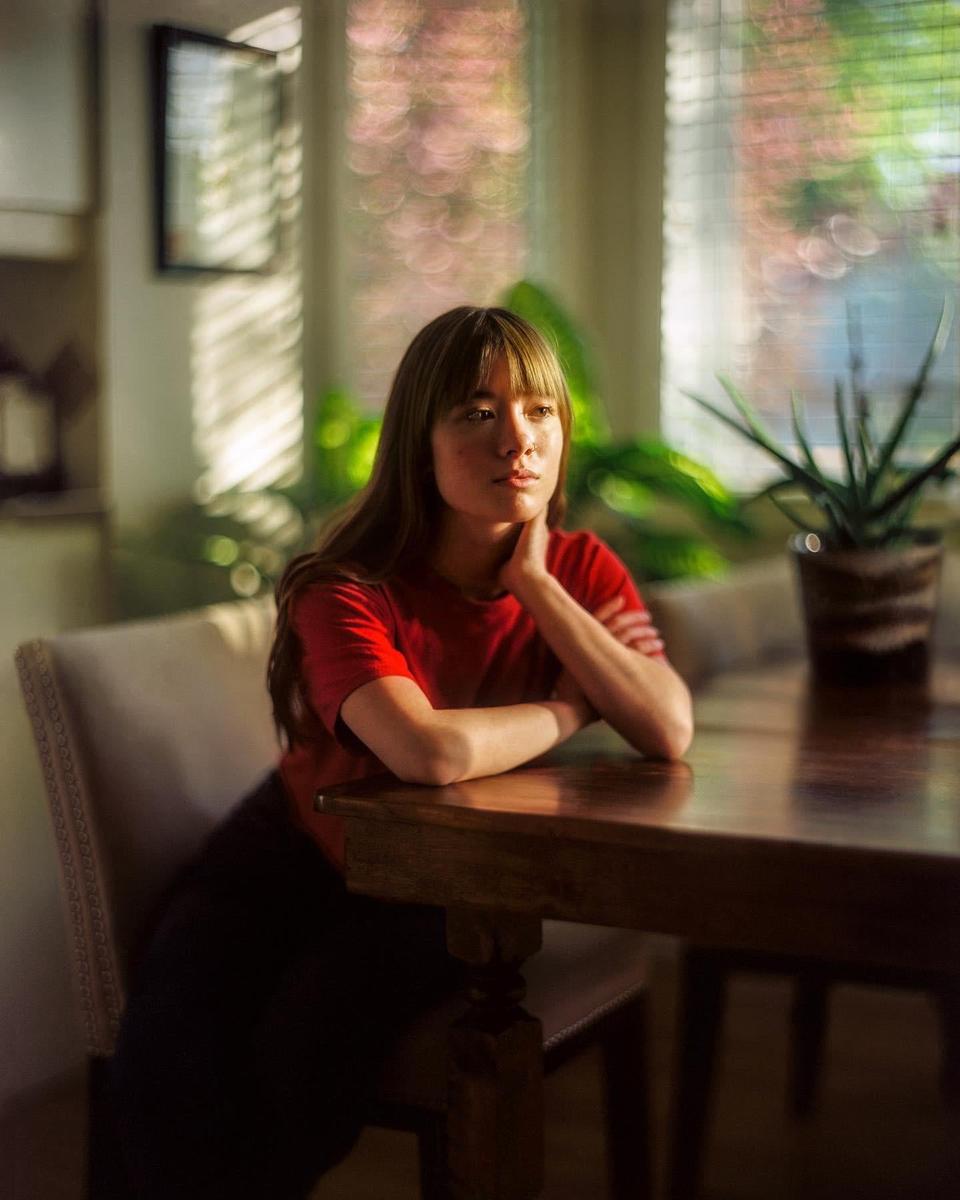 A woman in a red shirt sits at a wooden table with her arms crossed, looking out a window with light streaming through blinds.