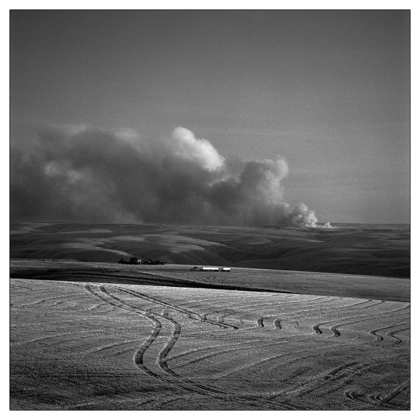 Smoke rises from a fire in the distance across a field with tire tracks in the foreground.