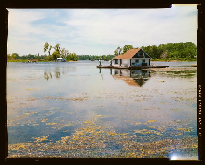 A small houseboat with a brown roof is floating on a lake with green algae and is reflected in the water.