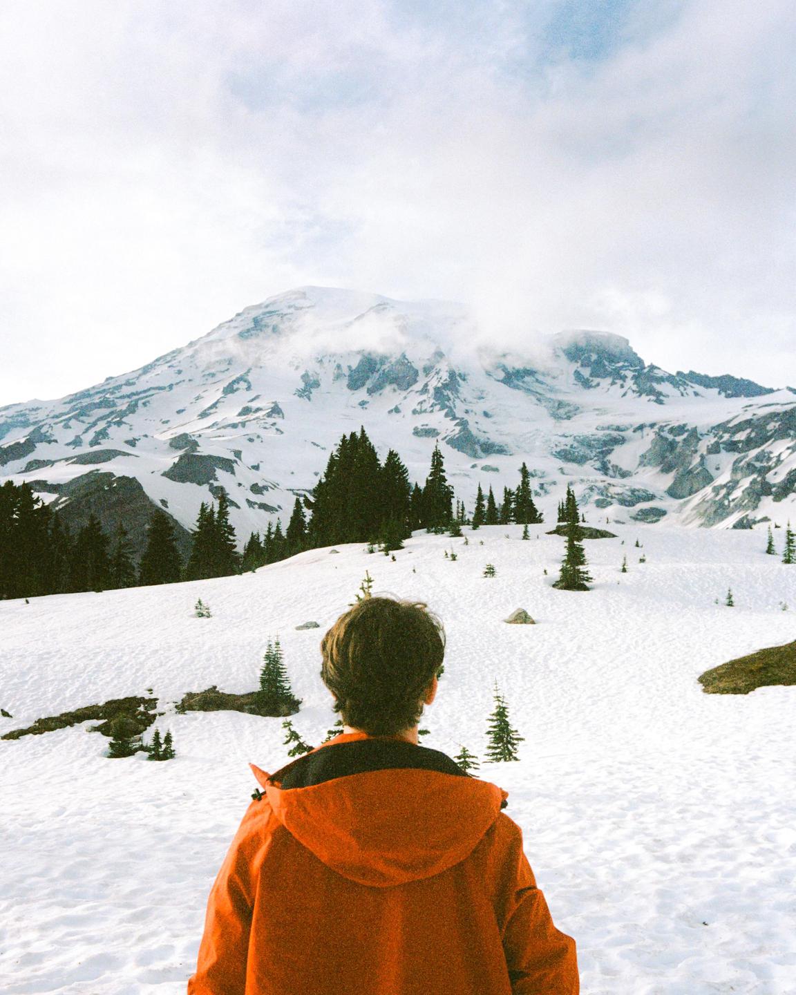 A person in an orange jacket stands in a snowy landscape looking at a large mountain.