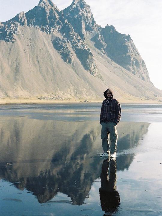 A man stands on a wet beach with a large mountain reflecting in the water behind him.