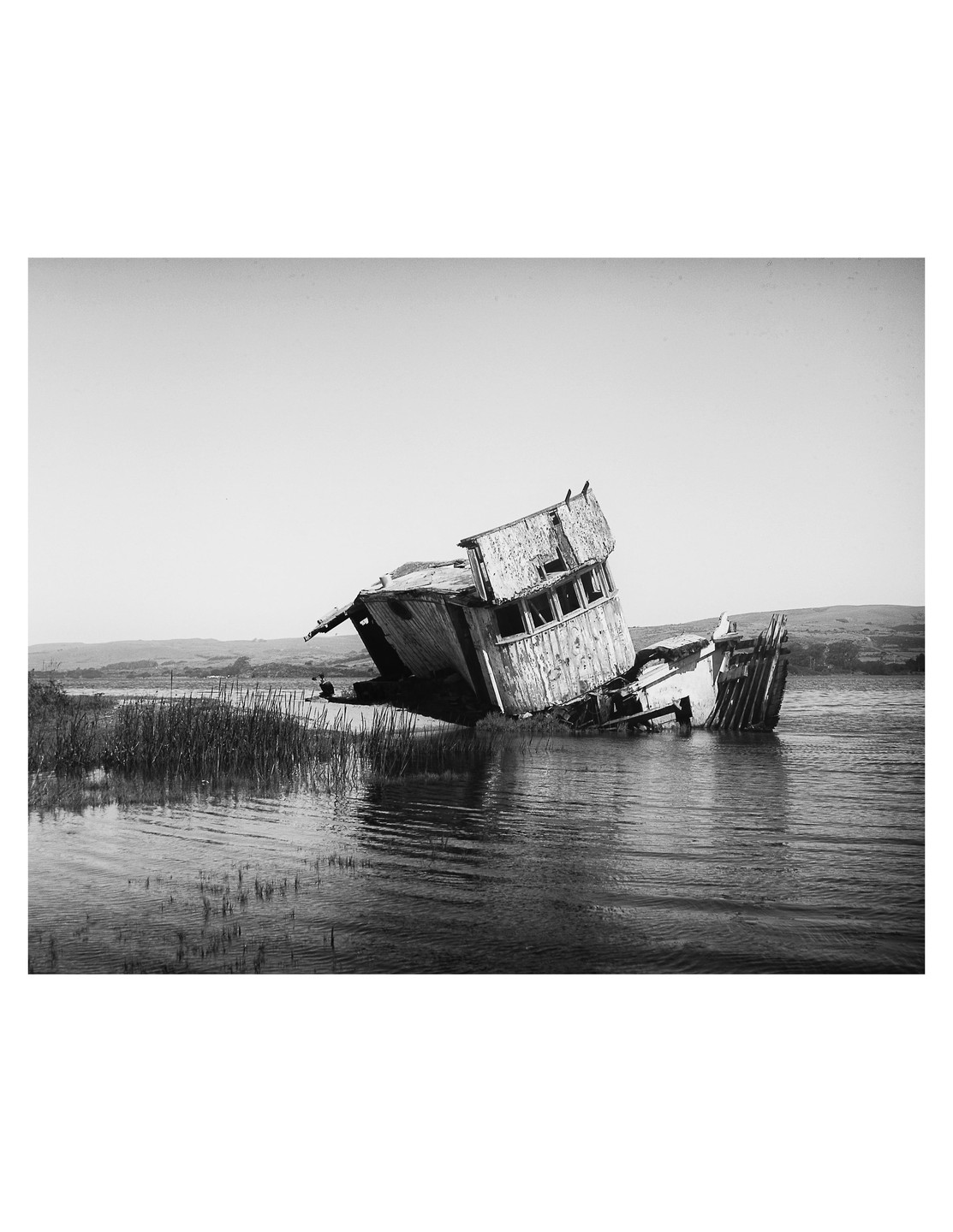 A decaying wooden shipwreck rests partially submerged in shallow water near a grassy shore.