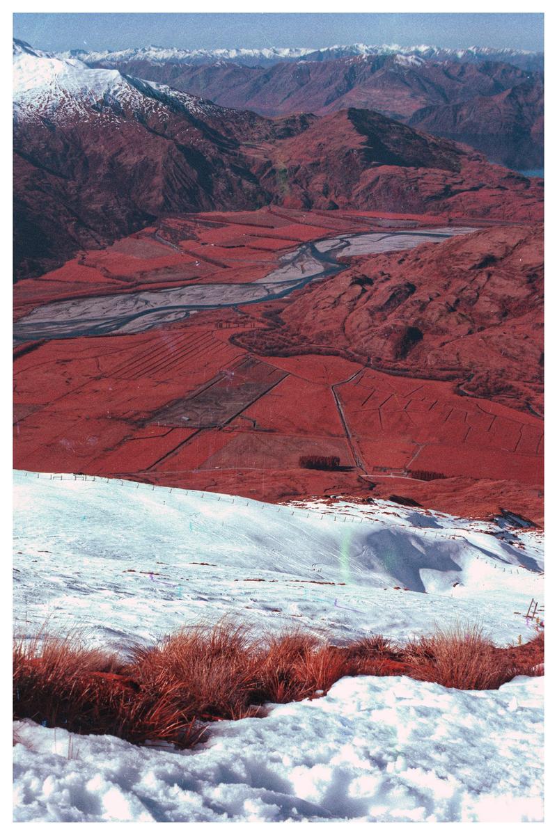 A snow-covered mountain slope with patches of dry grass overlooks a valley with a river and red-hued fields, surrounded by mountains.