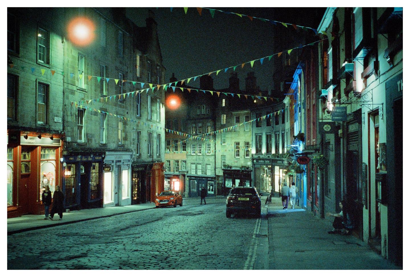 A cobblestone street in a city at night is lined with buildings and decorated with colorful bunting.