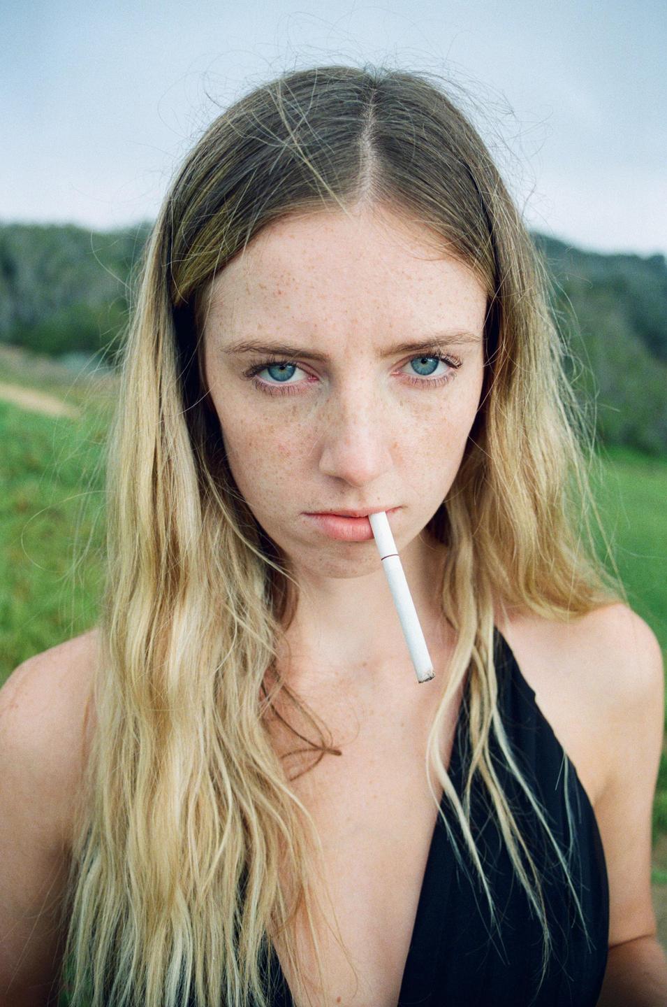 A woman with long blonde hair and freckles holds a cigarette in her mouth while looking at the viewer.
