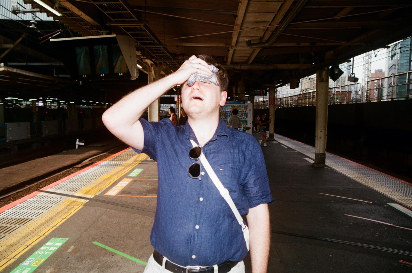 A man wipes his forehead with a cloth while standing on a train platform.