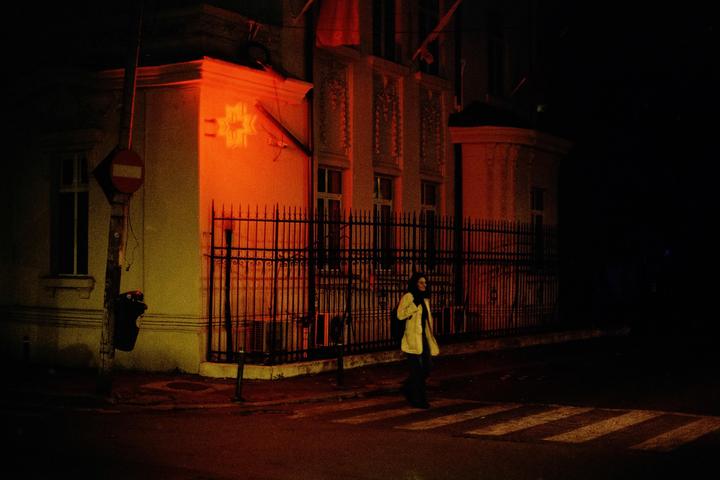 A woman crosses a street at night in front of a building with a fence and a red stop sign.