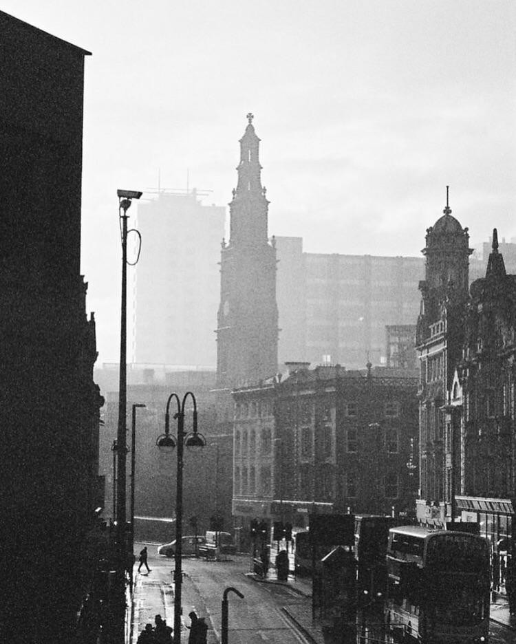 A street in Leeds is shown with a tall church steeple and other buildings in the background, and a street lamp and people in the foreground.