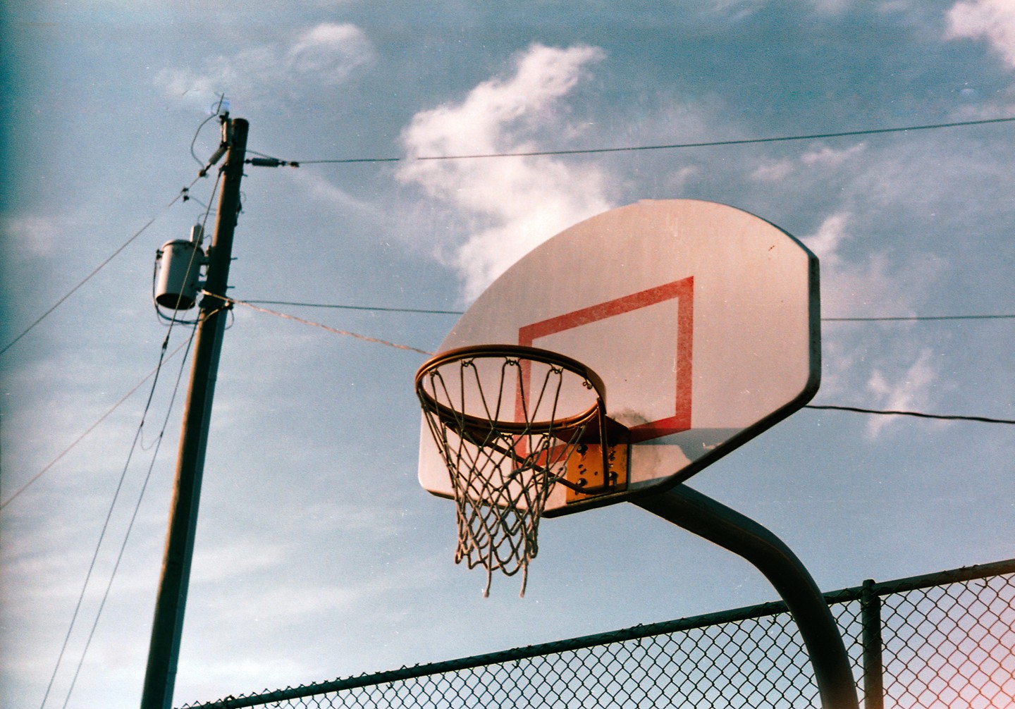 A basketball hoop and backboard are visible against a cloudy sky, with a utility pole and fence in the background.