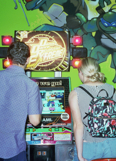 A man and a woman play an arcade game in a room with a Teenage Mutant Ninja Turtles mural.