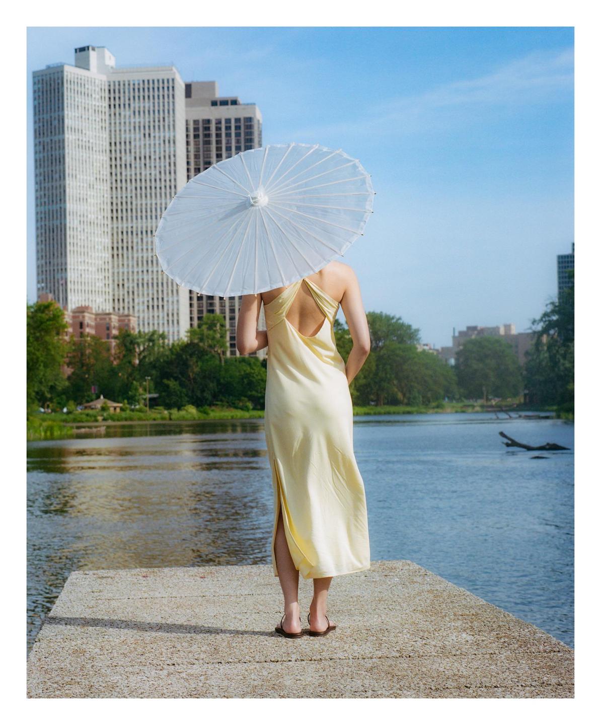 A woman in a yellow dress stands on a pier holding a white parasol, looking out at a body of water with city buildings in the distance.