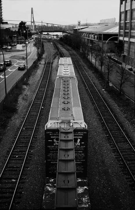 A freight train car is visible from above, with multiple train tracks, a road with cars, and a bridge in the background.