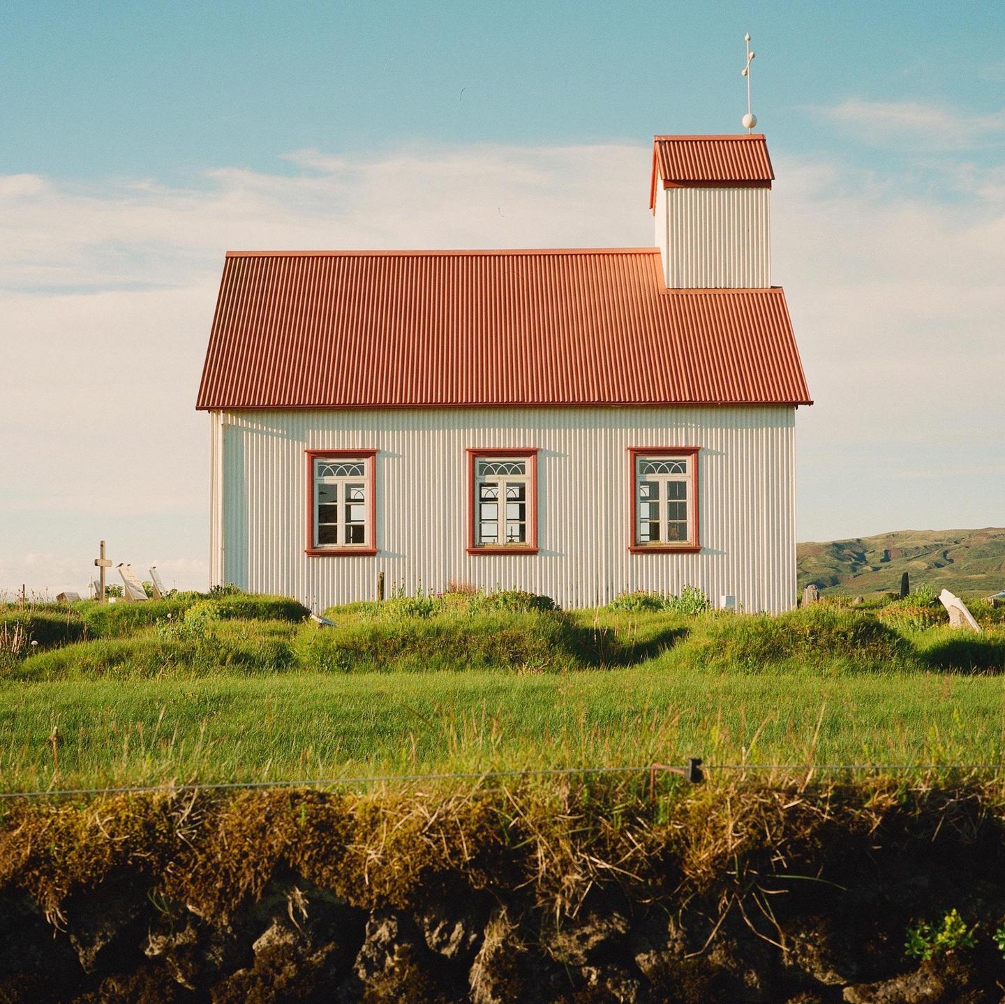 A white church with a red roof stands on a grassy hill under a blue sky.