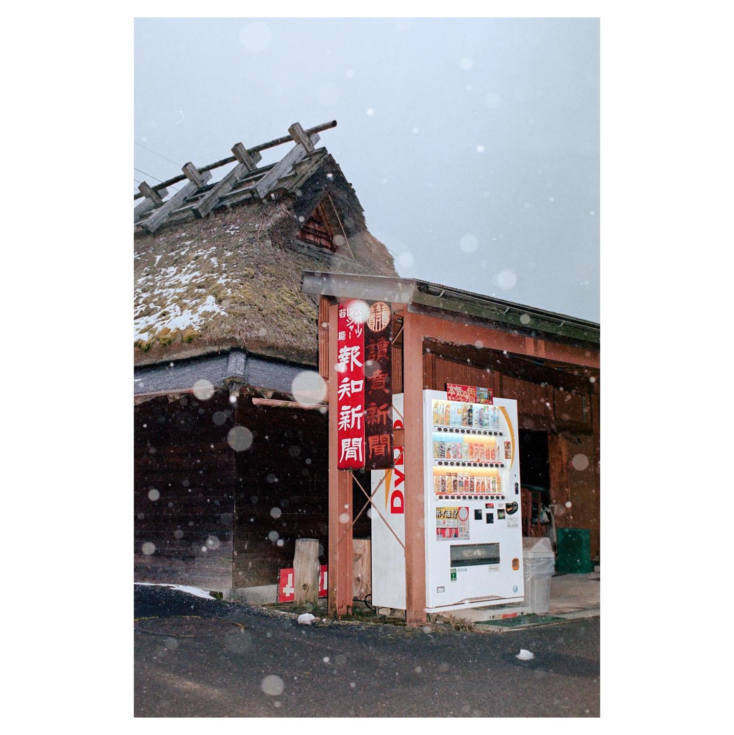 A traditional Japanese building with a thatched roof stands next to a modern vending machine as snow falls.