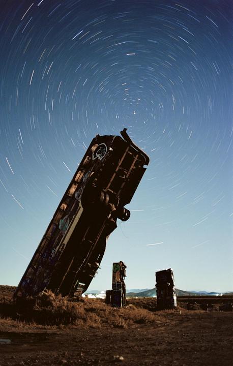 A car is buried nose-down in the ground with star trails visible in the night sky.