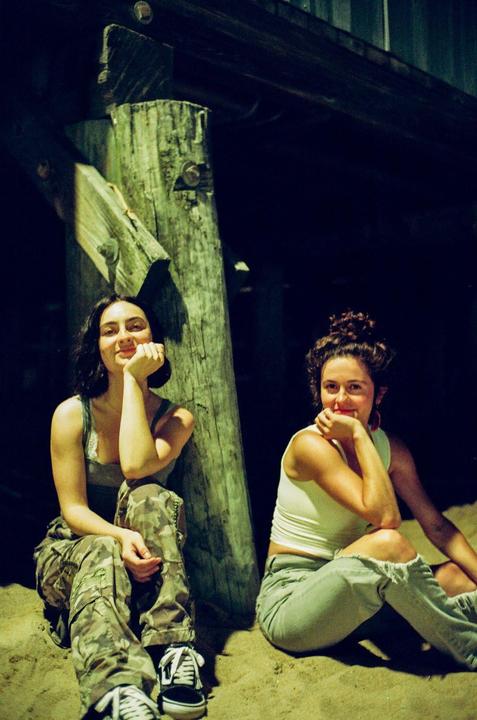Two women sit on sand under a pier, illuminated by a bright light source.