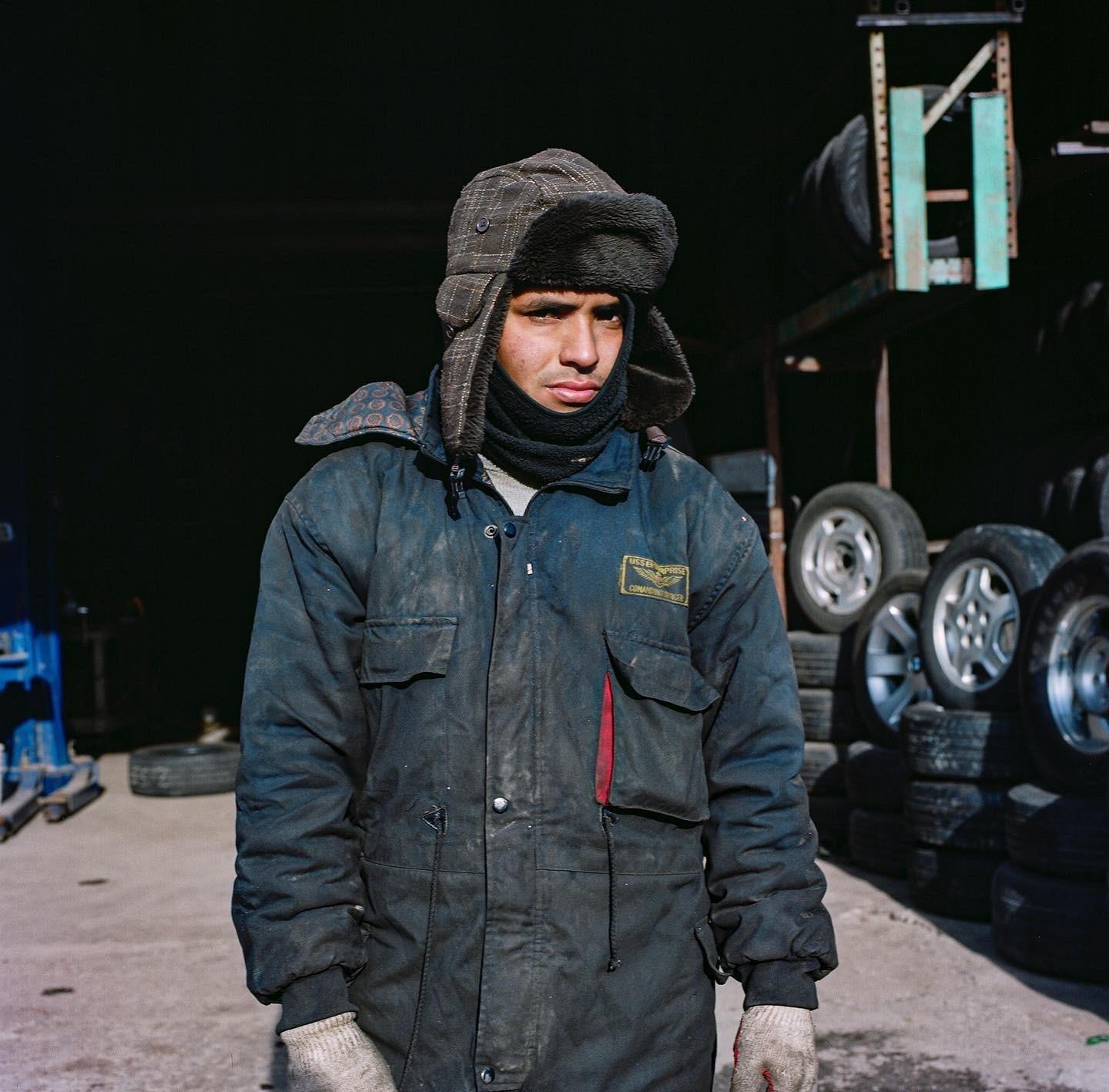 A man in a winter hat and work jacket stands in front of a stack of tires.