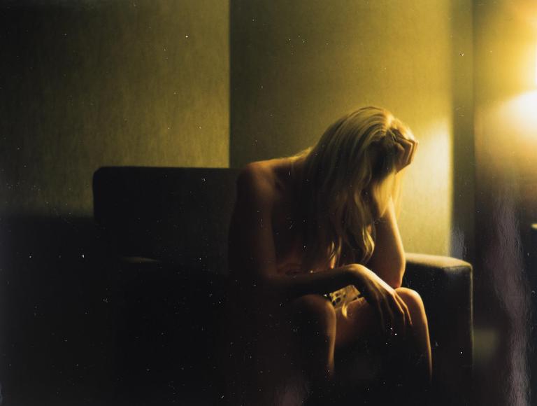 A woman with long blonde hair sits on a chair with her head in her hand in a dimly lit room.