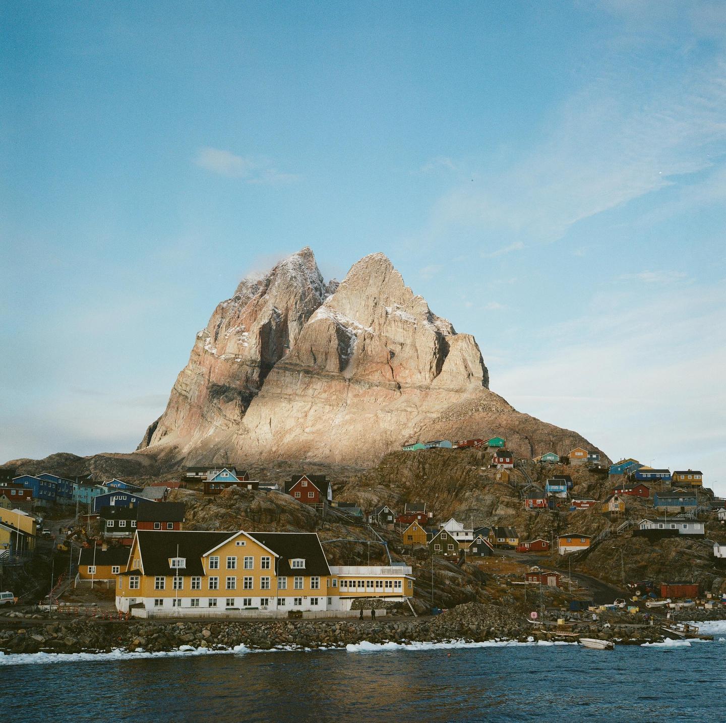 A small village with colorful houses is nestled at the base of a large, rocky mountain with snow-capped peaks, next to a body of water.
