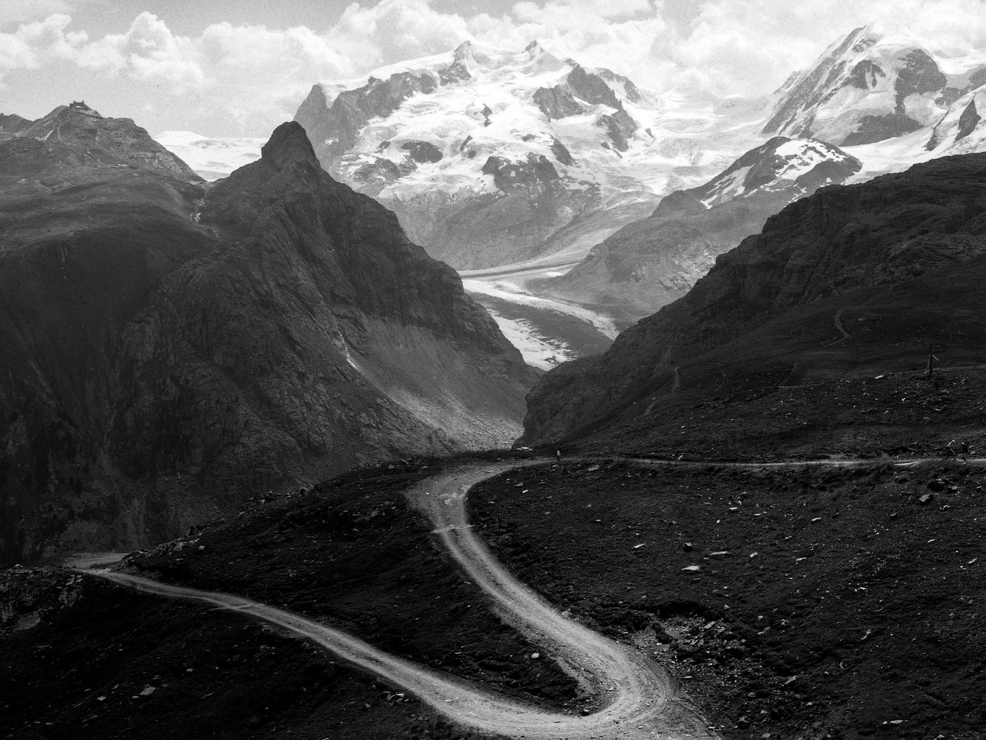 A winding dirt road leads through a valley towards snow-capped mountains and a glacier.