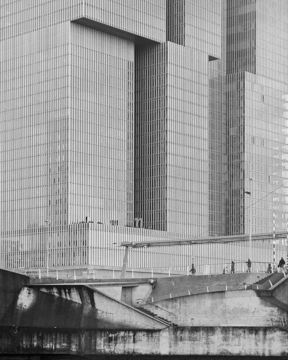 A large, modern building with many windows rises above a concrete structure with people walking on it.